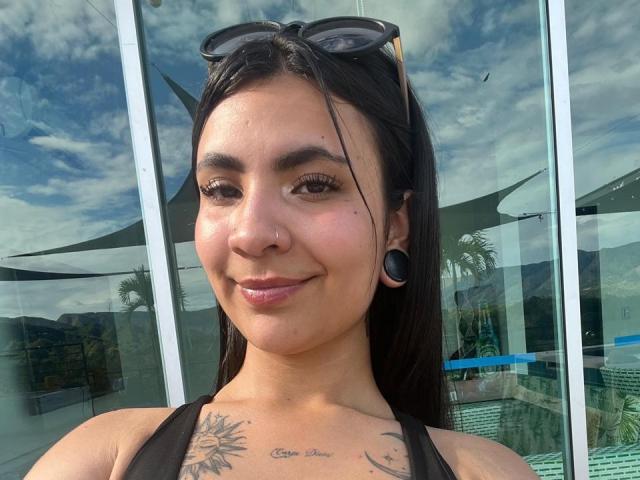 HanahCooper heeft donker haar, zwarte eyeliner, een neusring, zwarte oorbellen, een zonnebril op haar hoofd en tatoeages op haar borst, waarvan "Carpe Diem" zichtbaar is. Ze glimlacht met glanzende, roze lippen..