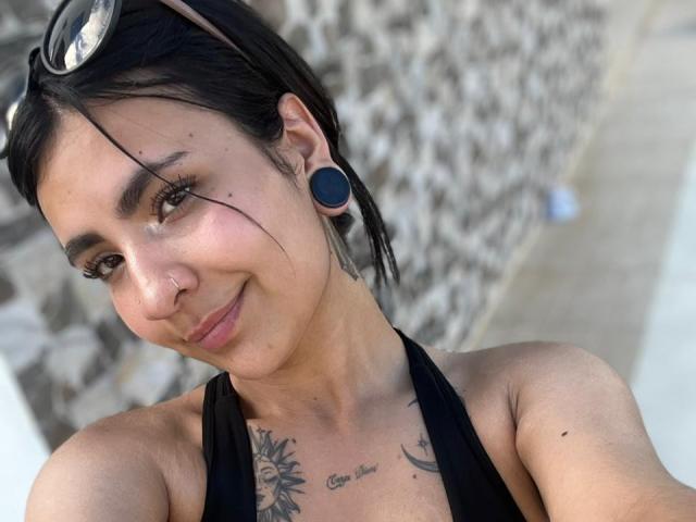HanahCooper with black ear gauges, nose ring, and small tattoo under her chin, wearing a black halter top, smiling slightly, dark hair, sunglasses on head, outdoor selfie.