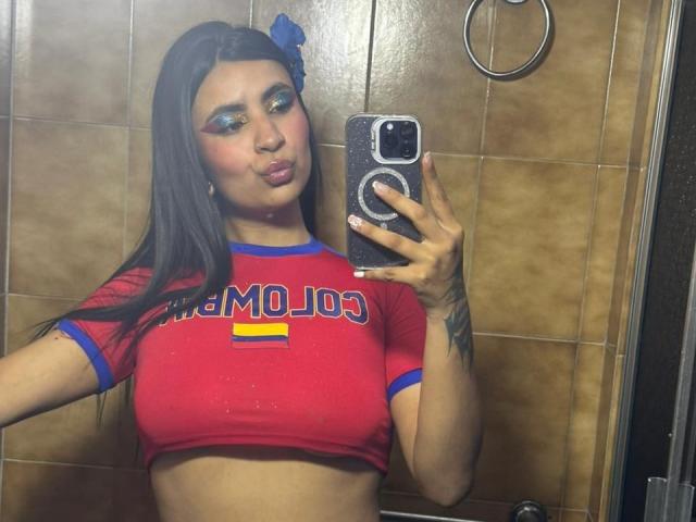 HanahCooper with big tits in a red crop top, pouting in a bathroom mirror selfie. She has dark hair, blue eyeshadow, and a blue headband. Her phone is in her right hand.