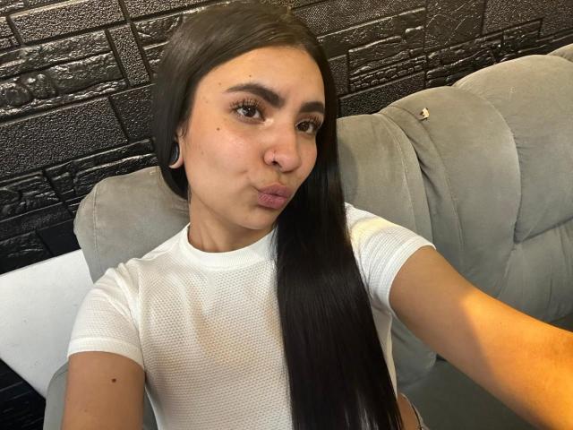 HanahCooper, com cabelos pretos longos, lábios cheios e uma blusa de malha branca, sentada em um sofá cinza, usando brincos grandes em formato de argola, olhando para a câmera com uma expressão levemente emburrada..