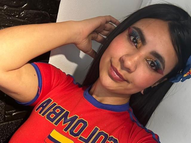 HanahCooper, avec ses cheveux foncés, son maquillage bleu pour les yeux et un anneau au nez, portant un maillot de foot rouge, souriant à la caméra, une main dans les cheveux, un accessoire bleu pour les cheveux..