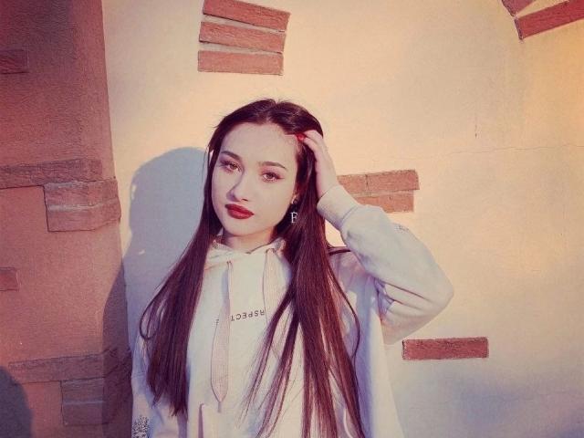Eleonora, con lunghi capelli scuri, indossa una felpa bianca larga, rossetto rosso, una mano tra i capelli, in piedi contro un muro di mattoni, guarda la fotocamera, illuminazione soffusa, stile casual..
