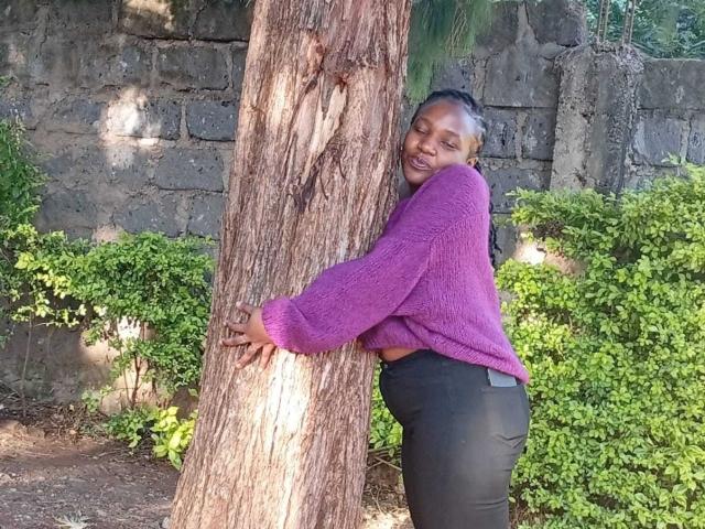 ClaireJulie hugging a tree with a purple sweater, black pants, and a smile. Her dark skin and braided hair are visible, and she's standing in front of a stone wall.
