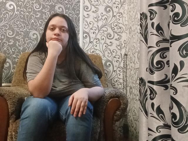 Leonore Gerrie, avec ses longs cheveux noirs, son t-shirt gris et son jean bleu, est assise sur un fauteuil marron et duveteux, une main sur son menton, et a l'air légèrement ennuyée. Ses fesses sont légèrement visibles..