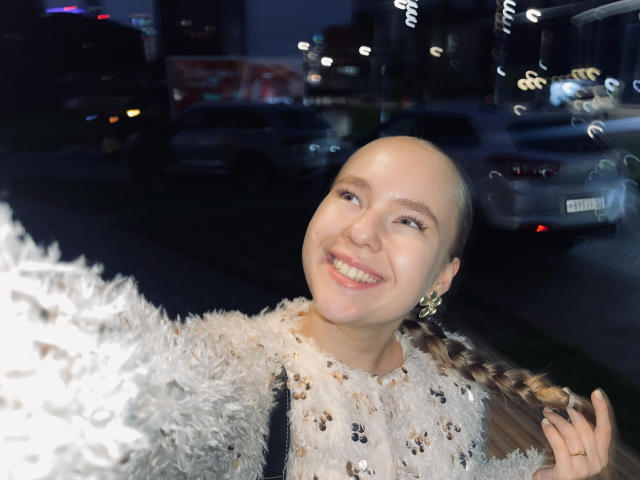 Karleen Carthen, con la cabeza rapada, sonriendo, vestida con una blusa blanca esponjosa, sosteniendo una trenza, de pie afuera por la noche, con luces de la ciudad al fondo, un coche plateado a la derecha..