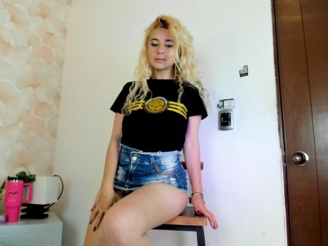 Sophie Teller, con i capelli biondi, maglietta nera con strisce gialle, pantaloncini di jeans, seduta su uno sgabello, gambe incrociate, mano destra sullo sgabello, mano sinistra sul ginocchio, aspetto casual, sicura di sé, sexy..