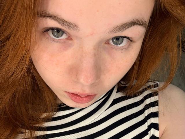 KatiRokisky's face with red hair, gray eyes, and freckles, wearing a black-and-white striped sleeveless top, looking up with slightly parted pink lips.