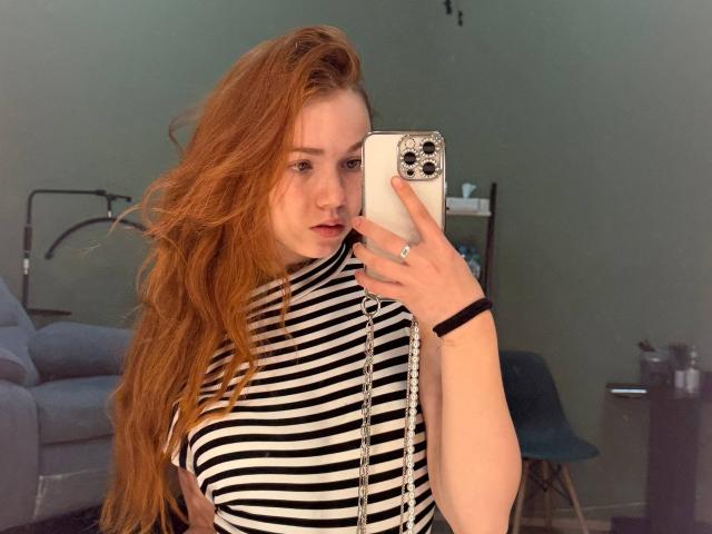 KatiRokisky, con lunghi capelli rossi, indossa una camicia a righe nere e bianche, tiene in mano un iPhone bianco, sta scattando un selfie allo specchio, porta un braccialetto nero ed ha un'espressione leggermente infastidita..