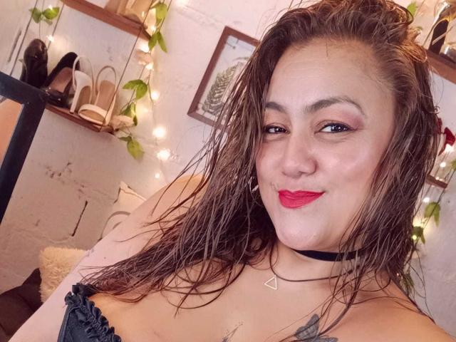 MissRosario, avec ses cheveux bruns mouillés, son rouge à lèvres rouge, son collier noir et son pendentif triangulaire, sourit à l'appareil photo, montrant ses petites seins et son tatouage sur sa poitrine droite..