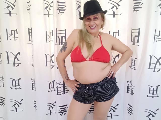 MatureYayita with blonde hair, black hat, red bra, showing her tits, and black sparkly shorts, smiling, hands on hips, tattoo on upper arm.
