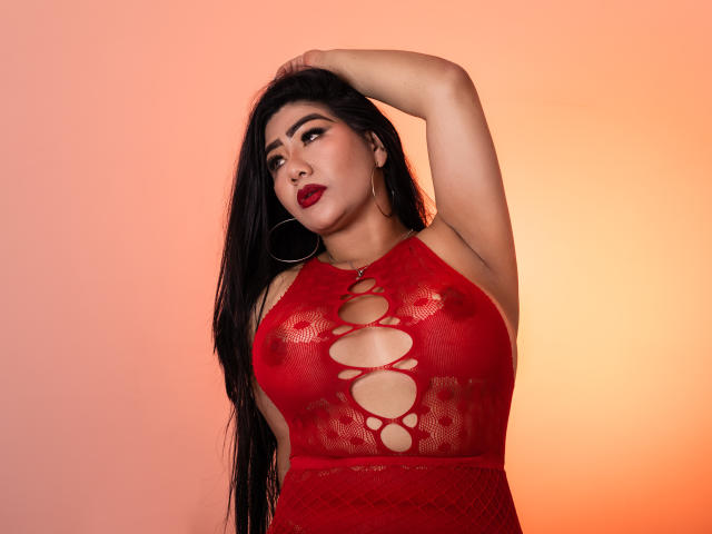 Khloe Lawson, con cabello negro largo, top de malla roja, busto grande y aberturas para los pezones, mano en el cabello, lápiz labial rojo, aros grandes, sobre un fondo degradado naranja..