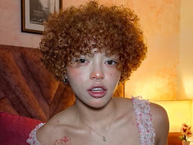 KeiryGinger has curly red hair, a nose ring, and is looking at the camera with pink lipstick and a small pimple on her nose. She's wearing a lacy white top.