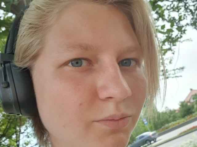 Das Gesicht von Laila Lafreniere, mit schwarzen Kopfhörern, kurzen blonden Haaren, blauen Augen und einem leichten Schmollmund, zeigt ein paar kleine Pickel auf ihrer Nase..
