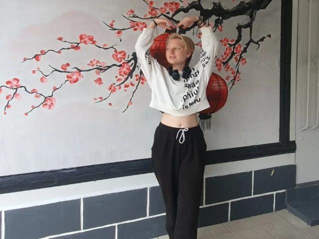 Laila Lafreniere, con cabello rubio, brazos levantados, vestida con un top corto blanco y pantalones deportivos negros, de pie frente a una pared decorada con un farol rojo y motivos de flores de cerezo..
