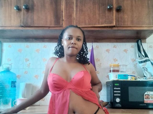 Yamila, com cabelos escuros e cacheados, usando um top de lingerie vermelho, expondo seus pequenos seios e calcinha preta, está em uma cozinha com armários de madeira e um micro-ondas..
