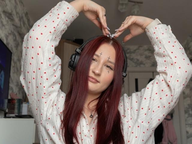 Tameika Vannoy, con i capelli rossi, che indossa delle cuffie, una camicia bianca a pois, con le braccia alzate, piccoli tatuaggi a forma di croce sotto gli occhi, sorride dolcemente..