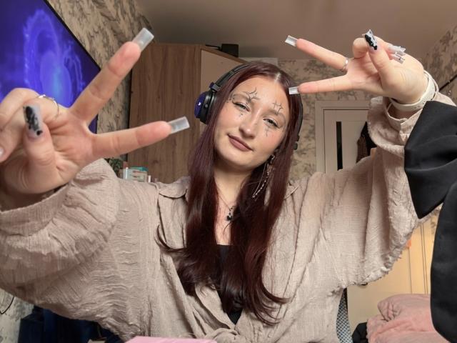 Tameika Vannoy, com cabelos castanhos longos, usando fones de ouvido, camisa bege, unhas pretas, tatuagens de estrelas, fazendo o sinal de "paz" com as duas mãos, sorrindo e exibindo suas unhas acrílicas brancas..