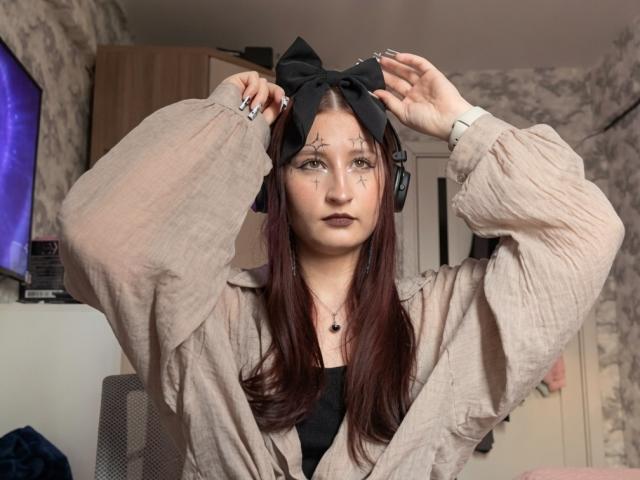 TameikaVannoy with red hair, black bow, and tattooed face, wearing a beige shirt, black top, and headphones, adjusting her bow with hands up.