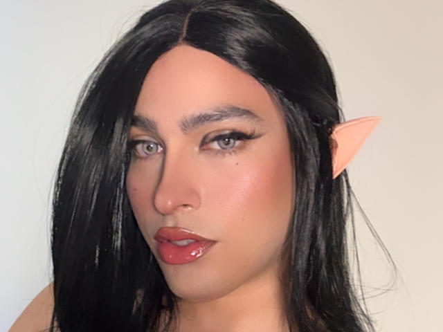 NaomiMoore69 a les cheveux noirs, des oreilles d'elfe de couleur rose pâle, des yeux bleus et des lèvres rouges brillantes. Son visage est légèrement rougi, et elle regarde la caméra avec assurance..
