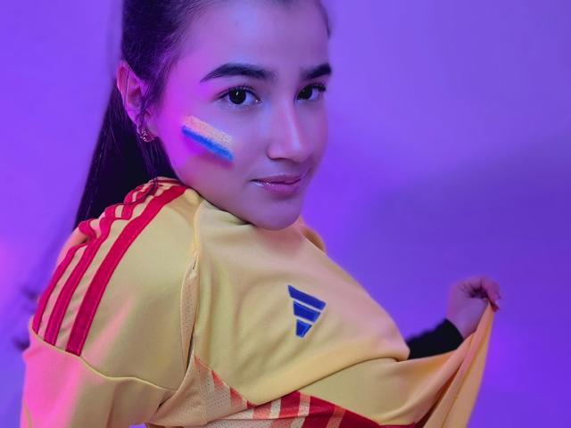 IrisBlair with blue and yellow face paint, yellow sports jersey with red stripes, and purple lighting, holding jersey, showing off tits. Long dark hair, cute, confident expression.