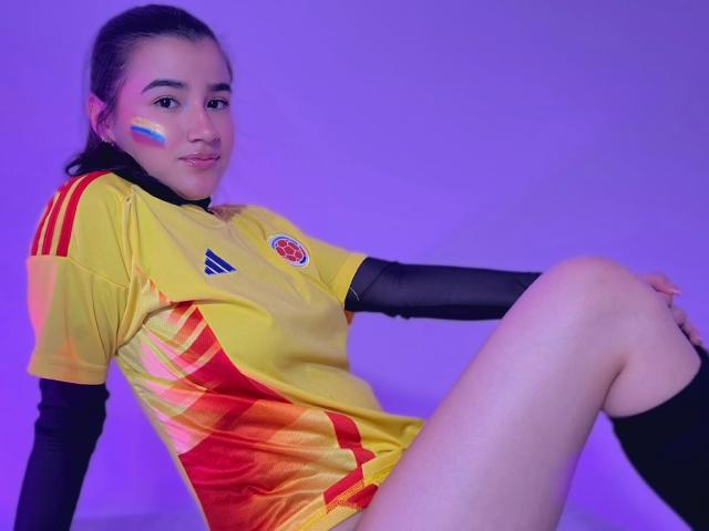 IrisBlair with yellow and red soccer jersey, black sleeves, and white face paint, sitting with legs spread, showing her ass, and a cute smile, in purple lighting.