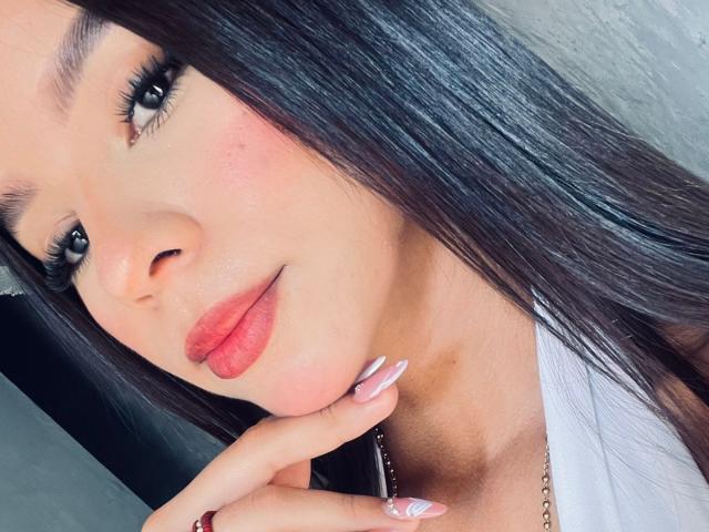 Le visage de Victoria Novak, avec ses cheveux noirs brillants, son rouge à lèvres rose et ses ongles blancs ornés d'un motif rose subtil. Elle touche son menton avec son doigt et sourit légèrement..