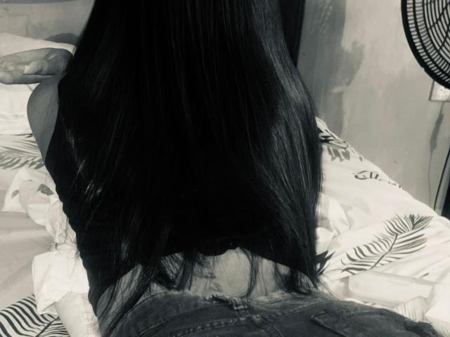 VictoriaNovak's long black hair, back to the camera, lying face down on a bed with her ass up, wearing jeans, in a grayscale photo.