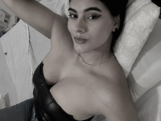 AlaraKey, con un seno prosperoso, sdraiata su un letto, indossa una canottiera di pelle nera, eyeliner nero, una collana a catena, capelli scuri e un'espressione leggermente accigliata, in una fotografia in bianco e nero..