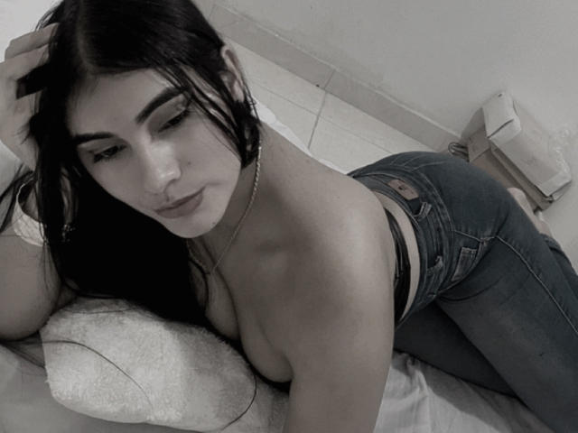 AlaraKey, con el pelo largo y negro, está acostada sin sostén en una cama, vestida con jeans azules, mostrando su trasero, mirando a la cámara con una ligera sonrisa, con una mano en el pelo..