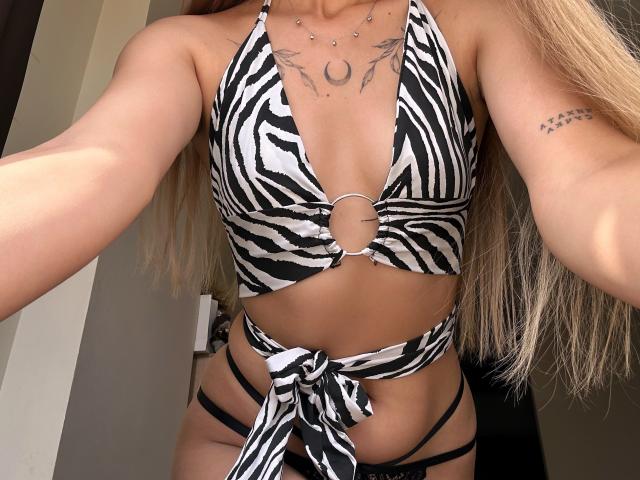 Karish, en maillot de bain rayé noir et blanc, montrant ses seins, son nombril percé et un bas noir à fines bretelles. Tatouage sur la poitrine, longs cheveux blonds, tatouage sur le bras avec des étoiles. Autoportrait..