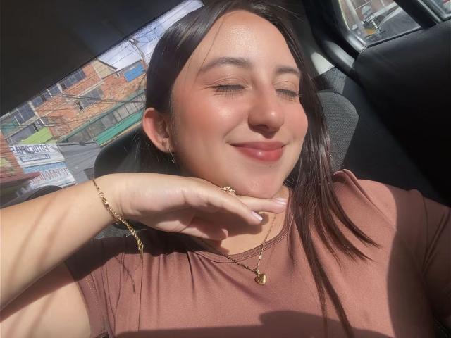 RosalieDupont, sonriendo con los ojos cerrados, rostro iluminado por el sol, camisa marrón, collar dorado, pulsera, mano en la mejilla, cabello marrón oscuro, interior de un coche, luz suave, relajada, linda..