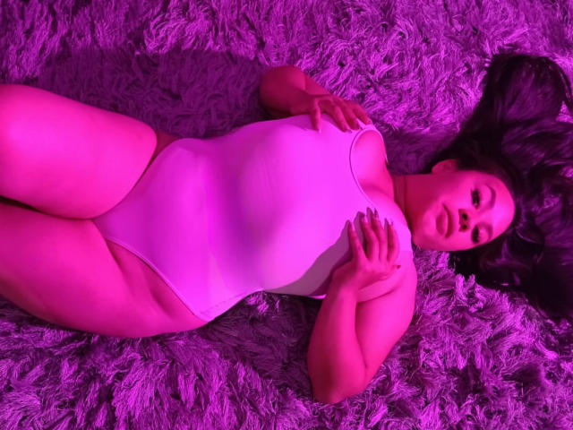 AshleyStart liegt auf einem violetten, flauschigen Teppich und trägt einen weißen Einteiler, der ihr großes Gesäß und ihre Brüste zeigt. Ihre Hände liegen auf ihrer Brust, und die Szene ist in rosa Licht getaucht..