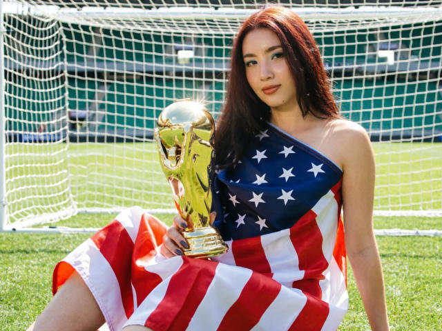 StarDustt, avec des cheveux roux, portant une robe aux couleurs du drapeau américain, tenant un trophée de football doré, assise sur de l'herbe verte, devant un but de football. Les seins sont partiellement visibles..