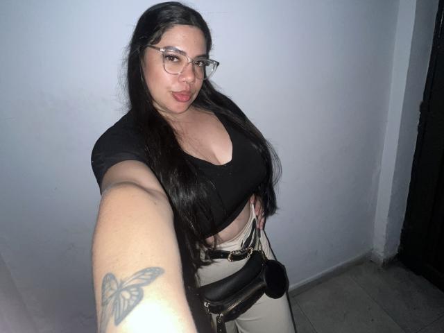 AndreaBusty, avec de longs cheveux noirs, des lunettes, un crop top noir, montrant de grandes seins, un tatouage sur le bras, tenant un sac, selfie, mur bleu pâle, sol carrelé et porte sombre en arrière-plan..