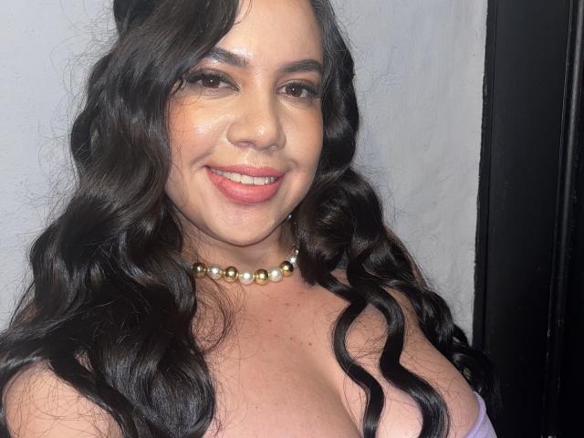 Andrea Busty, com longos cabelos negros ondulados, sorrindo, usando um colar de pérolas e exibindo seus seios grandes em uma blusa com decote baixo, tem pele clara e lábios rosados cheios..