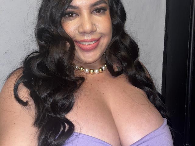 AndreaBusty tiene senos grandes y voluptuosos, lleva un top púrpura sin tirantes, con cabello negro largo y ondulado, un collar de perlas y una sonrisa encantadora, sobre un fondo sencillo..