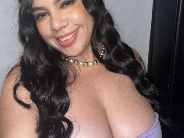 Andrea Busty mit großen Brüsten, lächelnd, mit langen, welligen schwarzen Haaren, die eine Perlenkette trägt und ein violettes Top ohne Träger, das ihre Brust zeigt. Ihre Haut ist hell und sie ist süß..