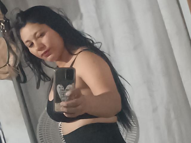 OliveTavy, avec ses longs cheveux noirs, portant un soutien-gorge de sport noir, prend un selfie avec son téléphone, montrant ses seins, dans une pièce avec un rideau blanc et une chaise..