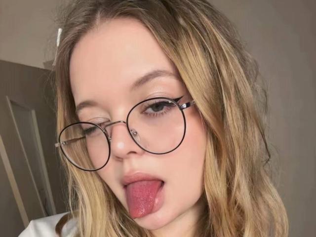 TanaHard, con cabello rubio y ondulado, gafas redondas y un piercing en la nariz, sacando su lengua rosa. Tiene piel clara y maquillaje ligero..