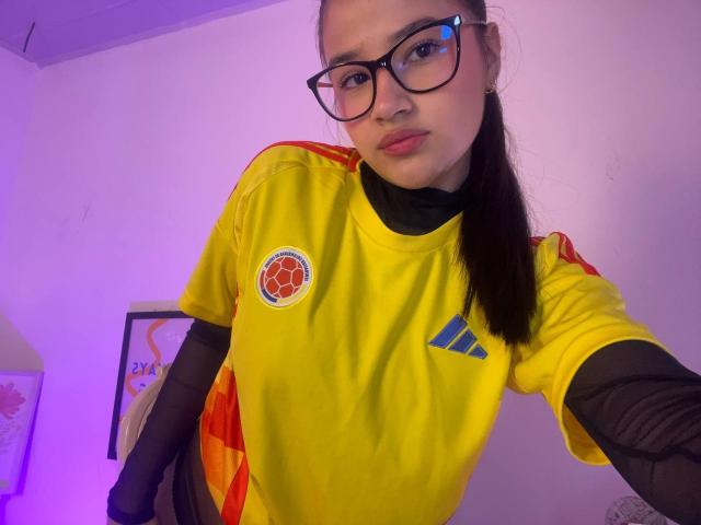 IrisBlair with glasses, wearing a yellow soccer shirt with red and blue accents, black sleeves, and a black turtleneck, leaning forward in a purple-lit room.