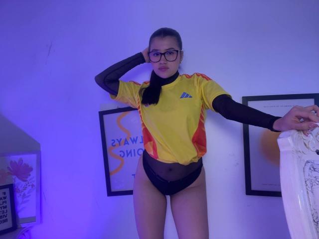 IrisBlair with glasses, yellow and red soccer shirt, black mesh long sleeves, black lace panties, standing, hand on head, blue-purple light, straight brown hair, white wall.