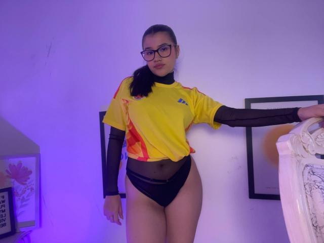 IrisBlair with glasses, yellow sports top, black mesh sleeves, black high-cut panties, standing, right arm on white bed, left arm raised, purple room lighting.