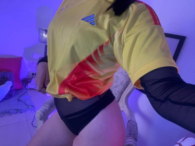 IrisBlair in a yellow and red soccer jersey, black mesh sleeves, and black lace panties, showing her thighs and ass, with blue lighting in a room.