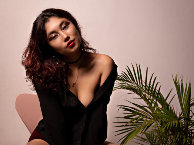 AliCipher with red lipstick, wavy dark hair, black off-shoulder shirt, and necklace, leaning forward, showing her small tits, green plant to the right, pink chair in background.