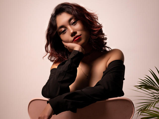 AliCipher with wavy dark red hair, wearing an off-shoulder black shirt, resting her chin on her hand, showing her tits, sitting on a pink chair, red lipstick.