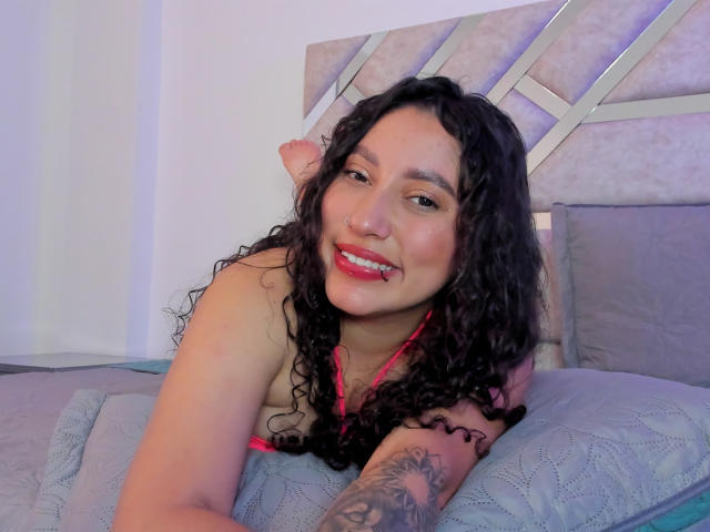 AmaraDeLux with curly black hair, pink bra, and tattoo on right arm, smiling on bed, pink lipstick, lying on grey pillows, showing her tits.