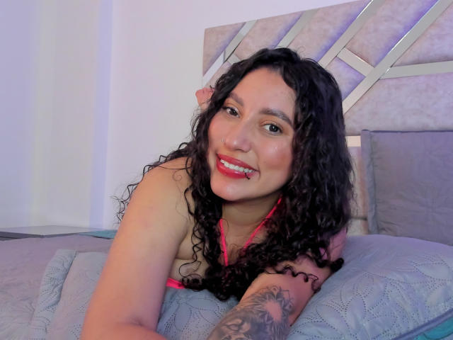 AmaraDeLux with curly black hair, red lipstick, and pink bra, smiling, lying on bed, showing tattoo on right arm, white skin, and pink nose ring.