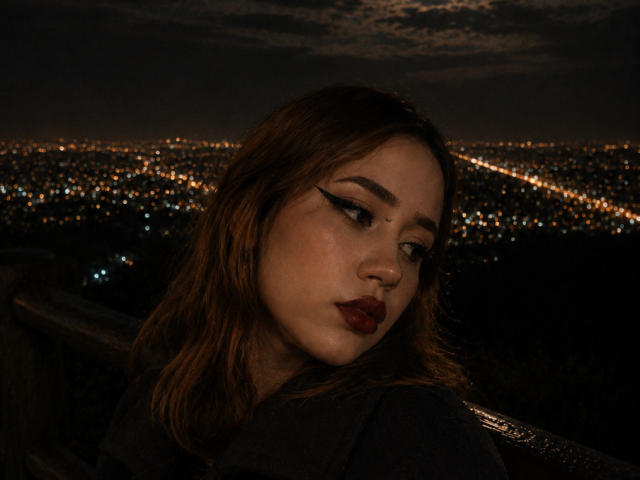 EmyHouston a les cheveux brun foncé, un trait d'eye-liner épais, du rouge à lèvres rouge et une expression légèrement boudeuse. Elle porte une chemise sombre et regarde sur le côté, dans un paysage urbain nocturne..