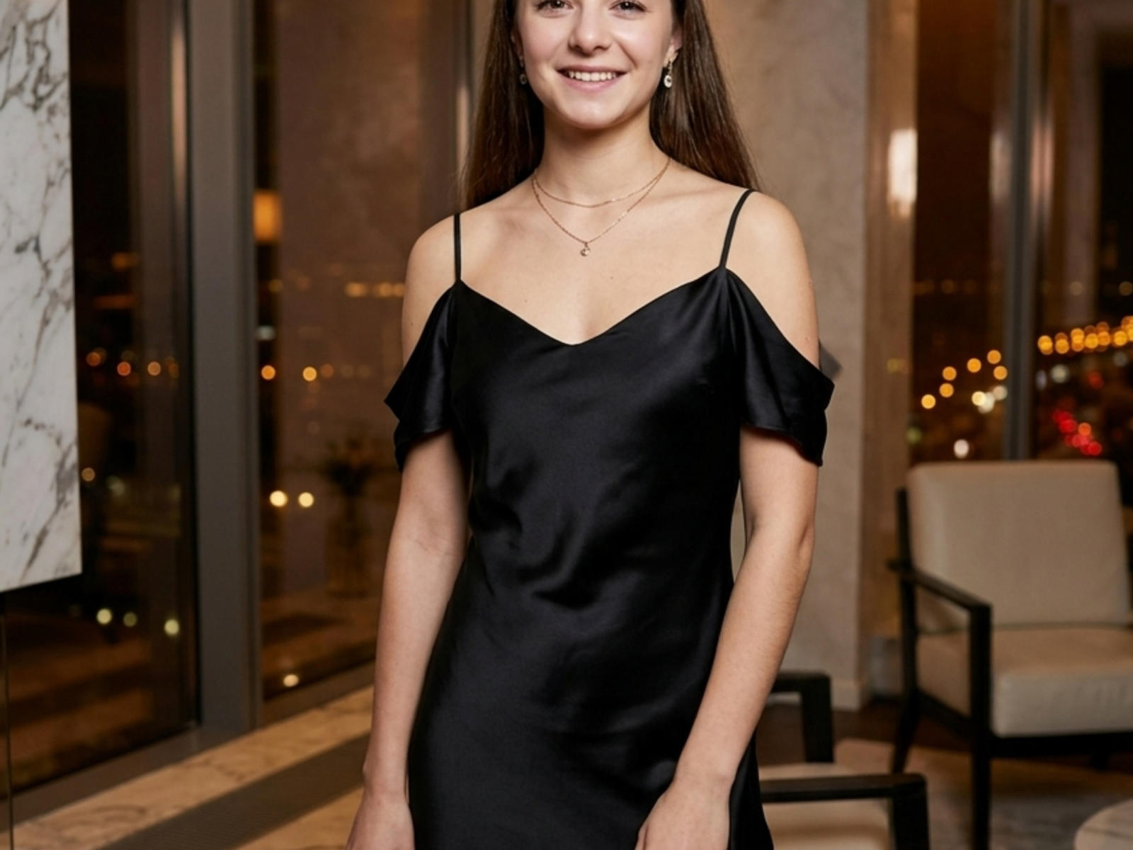 Marianela Walstrom, vêtue d'une robe en satin noir à une épaule, souriante, avec de longs cheveux bruns, portant un collier, se tient à l'intérieur avec une vue nocturne de la ville à travers des fenêtres en verre..