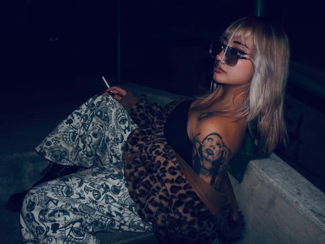 AgathaLin met blond haar, een zonnebril en een jas met luipaardprint, zittend op beton, een sigaret vasthoudend, met tatoeages op haar borst en arm, en gekleed in broek met een patroon..